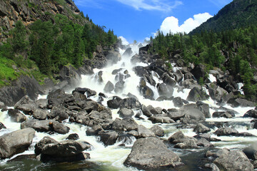 Wall Mural - A powerful stream of water from the Uchar waterfall rushes over huge stones from the mountain, coniferous trees grow on the rocky slopes of the mountains, a clear sunny day, the sky with clouds