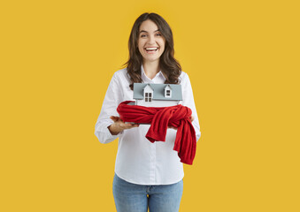 Joyful woman holds toy house wrapped in warm scarf as symbol for heating system or cold snowy winter. Isolated on orange background. Building insulation, heating season, warm up and protect concept.