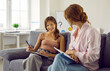 © Studio Romantic - Therapist working with teenage child. Happy adolescent girl sitting on sofa with psychologist, holding pen and mental health questionnaire, honestly answering questions and discussing her answers