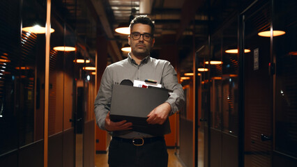 Fired upset man in suit walking down in office hallway, holding box with books and personal stuff, saying goodbye to colleagues, looking at camera. Male employee leaving marketing company