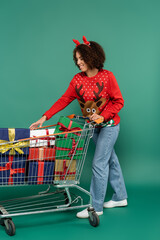 Wall Mural - african american woman in deer horns headband taking present from shopping cart on green background