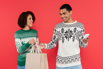 Wall Mural - smiling african american woman holding shopping bags near man with smartphone isolated on red