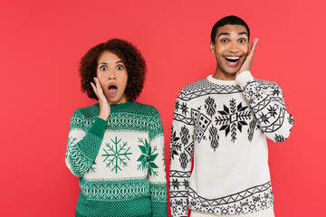 Wall Mural - thrilled african american couple in warm knitted sweaters looking at camera isolated on red