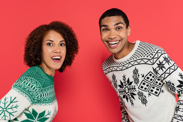 Wall Mural - excited african american couple in winter sweaters with ornament looking at camera isolated on red