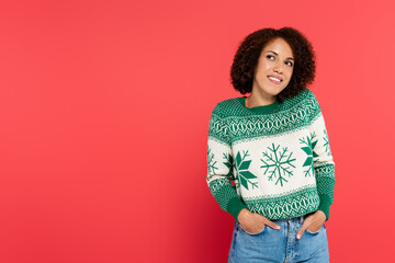 Wall Mural - dreamy and smiling african american woman in winter sweater posing with hands in pockets of jeans isolated on red