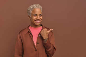 Wall Mural - Positive pretty African American woman in casual dressed smiling points to side and turns head to point to copy space for your advertisement for young girls stands on brown studio background