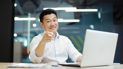 Asian office worker employee points a finger at the camera while sitting in office at workplace. Portrait of a handsome happy businessman in white shirt work laptop computer Looking at camera smile