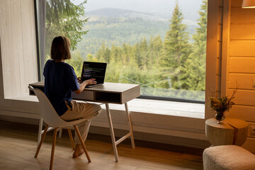 woman works on laptop while sitting by the table in front of panoramic window with great view on mou