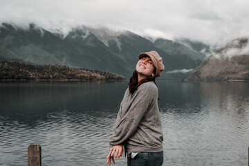 Canvas Print - happy caucasian girl smiling baring teeth wearing brown beanie gray sweatshirt with arms crossed and eyes closed enjoying calm lake in nature, nelson lakes, new zealand