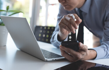 Wall Mural - Asian businessman working on laptop computer browsing the internet, using mobile smart phone with digital tablet on white office table, close up