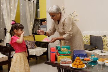 A grandmother offering sweets trying to please cute young little girl kid, dressed up in ethnic wear, celebrate diwali Hindu festival Laxmi poojan, kids expression