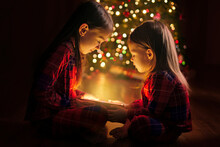 The Sisters Are Holding A Box With A Magical Glow From The Inside Sitting On The Floor By The Christmas Tree. Magic On Christmas And New Year's Eve. A Gift From Santa