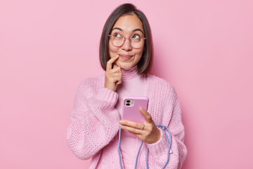 Studio shot of pretty Asian woman with dark hair keeps finger near lips has dreamy facial expression holds mobile phone looks aside wears spectacles and jumper isolated over pink background.