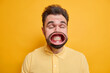 Portrait of dark haired man has widely opened mouth on strong wind shows white teeth dressed in shirt isolated over vivid yellow background. Crazy European male screams loudly stands indoor.