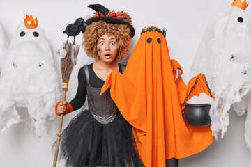 Wall Mural - Shocked scared woman pretends being witch embraces spooky ghost holds broom with crow celebrates Halloween at party stares amazed at camera cannot believe own eyes. Magic and witchcraft concept