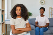 Attractive teenage girl is looking sadly away. Girl sits with her arms crossed in closed pose. Defocused Mom sitting behind and looks at her daughter. African american family at home.
