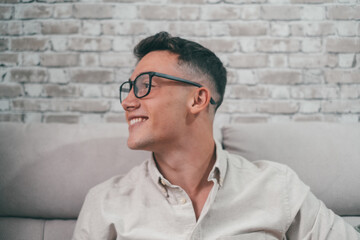 Portrait of one young attractive man looking at the window smiling and having fun sitting on the sofa. Happy male teenager wearing eyeglasses enjoying..