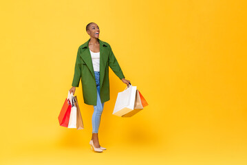 Wall Mural - Trendy fashionable African American woman carrying colorful shopping bags walking on yellow color studio isolated background