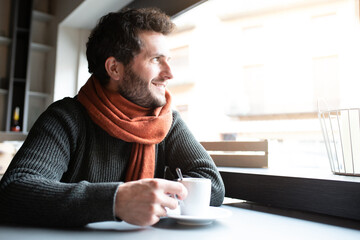 Pensive, happy young man drinking coffee in cafeteria looking out the window, enjoying early morning. Copy space.