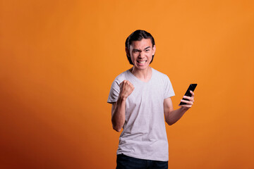 Young excited asian man showing winning gesture, good news celebration, victory concept. Joyful happy teenager with clenched fist holding smartphone, winner celebrating success