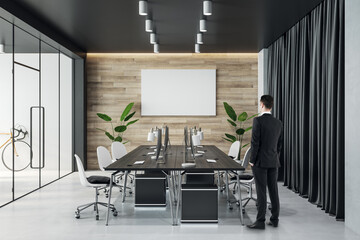 Canvas Print - Businessman back view looking at blank white poster with place for your logo or text on wooden wall in stylish board room with dark meeting table, white chairs and transparent doors, mock up