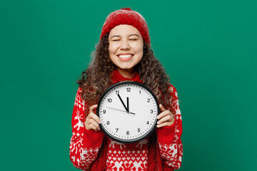 Merry young woman wearing red warm cozy knitted sweater hat posing hold clock waiting midnight isolated on plain dark green background studio portrait. Happy New Year 2023 celebration holiday concept.