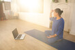 © Alberto Cotilla - The Yoga teacher dressed in blue is sitting on a mat while conversing with her students on a laptop in a video call, light is coming through the reeds on the wall.