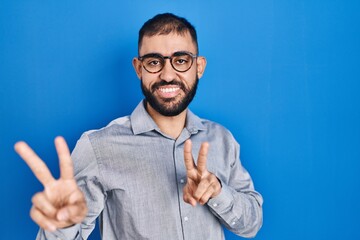 Wall Mural - Middle east man with beard standing over blue background smiling looking to the camera showing fingers doing victory sign. number two.