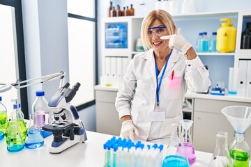 Wall Mural - Middle age blonde woman working at scientist laboratory pointing with hand finger to face and nose, smiling cheerful. beauty concept