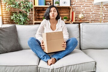 Wall Mural - Young hispanic woman sitting on the sofa checking delivery package looking at the camera blowing a kiss being lovely and sexy. love expression.