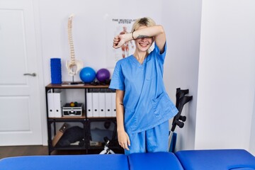 Wall Mural - Young physiotherapist woman working at pain recovery clinic covering eyes with arm smiling cheerful and funny. blind concept.