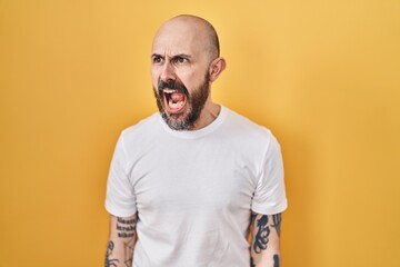 Poster - Young hispanic man with tattoos standing over yellow background angry and mad screaming frustrated and furious, shouting with anger. rage and aggressive concept.