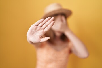 Poster - Young redhead woman standing over yellow background wearing summer hat covering eyes with hands and doing stop gesture with sad and fear expression. embarrassed and negative concept.