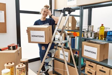 Canvas Print - Young caucasian woman volunteer holding donations box doing ok gesture with hand smiling, eye looking through fingers with happy face.