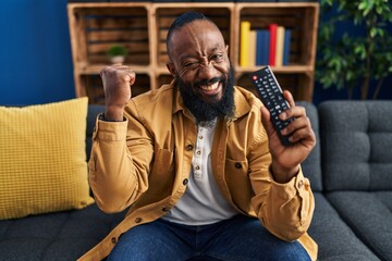 Poster - African american man holding television remote control screaming proud, celebrating victory and success very excited with raised arms