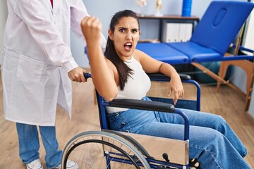 Wall Mural - Young hispanic woman sitting on wheelchair at physiotherapy clinic annoyed and frustrated shouting with anger, yelling crazy with anger and hand raised