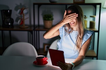 Sticker - Young hispanic woman using touchpad sitting on the table at night smiling and laughing with hand on face covering eyes for surprise. blind concept.