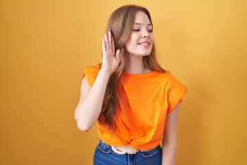 Poster - Caucasian woman standing over yellow background smiling with hand over ear listening an hearing to rumor or gossip. deafness concept.