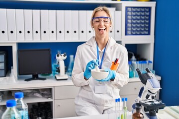 Poster - Young caucasian woman working at scientist laboratory sticking tongue out happy with funny expression.