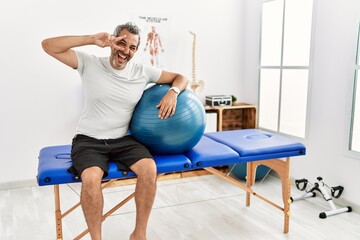 Wall Mural - Middle age hispanic man at pain recovery clinic holding pilates ball doing peace symbol with fingers over face, smiling cheerful showing victory