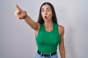 Sticker - Young woman standing over isolated background pointing with finger surprised ahead, open mouth amazed expression, something on the front