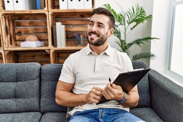 Canvas Print - Handsome hispanic man holding clipboard working at psychology clinic looking away to side with smile on face, natural expression. laughing confident.