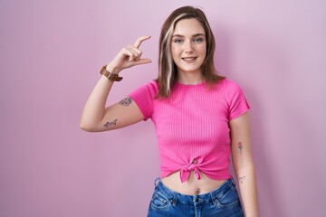 Sticker - Blonde caucasian woman standing over pink background smiling and confident gesturing with hand doing small size sign with fingers looking and the camera. measure concept.