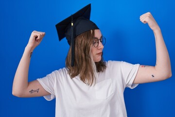 Poster - Blonde caucasian woman wearing graduation cap showing arms muscles smiling proud. fitness concept.