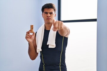 Sticker - Hispanic man eating protein bar as healthy energy snack pointing with finger to the camera and to you, confident gesture looking serious