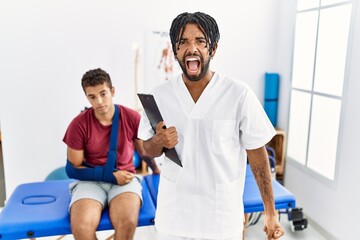 Canvas Print - Young hispanic man working at pain recovery clinic with a man with broken arm angry and mad screaming frustrated and furious, shouting with anger. rage and aggressive concept.