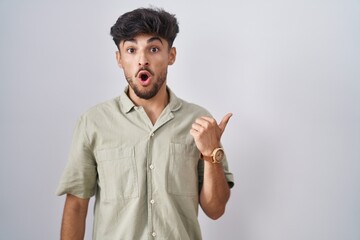 Sticker - Arab man with beard standing over white background surprised pointing with hand finger to the side, open mouth amazed expression.