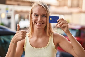 Sticker - Young caucasian woman holding credit card smiling happy and positive, thumb up doing excellent and approval sign