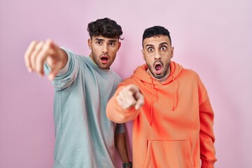 Wall Mural - Young hispanic gay couple standing over pink background pointing with finger surprised ahead, open mouth amazed expression, something on the front