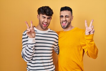 Wall Mural - Young hispanic gay couple standing over yellow background smiling with happy face winking at the camera doing victory sign with fingers. number two.